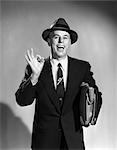 1950s SALESMAN MAN MAKING OKAY SIGN WITH THUMB AND INDEX FINGER CARRYING BRIEFCASE WEARING SUIT TIE AND HAT EAGER SMILING