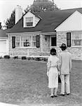 1940s 1950s COUPLE STANDING OUTSIDE LOOKING AT STONE SUBURBAN HOUSE