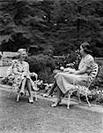 1940s TWO WOMEN SITTING IN LAWN CHAIRS TALKING SMILING BACKYARD GARDEN BOTH WEARING COTTON PRINT DRESS AND SPECTATOR SHOES