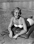 1940s SMILING BLOND WOMAN WEARING WHITE TWO PIECE BATHING SUIT LAYING ON INFLATED RAFT