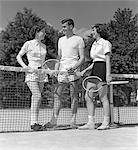 IN DER NÄHE 40ER JAHRE 50ER JAHRE VON ZWEI MÄDCHEN UND EIN KERL-TENNIS-SPIELER HOLDING TENNISSCHLÄGER UND TENNISBALL STANDING NET ON COURT