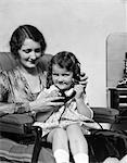 1920s 1930s SMILING GIRL SITTING ON MOTHERS LAP IN CHAIR TALKING ON TELEPHONE WITH MOTHERS HELP HOLDING RECEIVER
