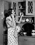 1920s 1930s WOMAN HOUSEWIFE WEARING APRON IN KITCHEN TALKING ON TELEPHONE PUTTING CANISTER ON CUPBOARD SHELF