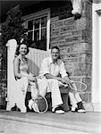 ANNÉES 1930 L'HOMME ET LA FEMME ASSISE SUR PORCHE TENUE TENNIS RAQUETTES SOURIANT L'ÉTÉ EN PLEIN AIR