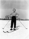 ANNÉES 1940 SOURIANTE BLONDE FEMME ON SKIS DEUX HANCHES ON DE MAINS TENANT DES BÂTONS DE SKI DANS LA NEIGE CHAMP AVEC ARBRE ARRIÈRE-PLAN MITAINES