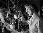 1950s SMILING PRETTY YOUNG WOMAN HANGING GLASS CHRISTMAS ORNAMENT ON BRANCH LIGHT COMES FROM THE TREE