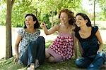 Three young women sitting in park, laughing, one pointing