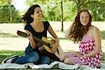 Two young women sitting on blanket outdoors, one playing acoustic guitar