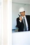 Man in suit, wearing hard hat, using walkie talkie, looking out window