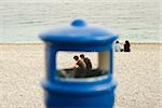 Couple sitting on the beach viewed through a garbage bin, Baie Des Anges, Nice, Provence-Alpes-Cote D'Azur, France