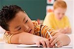 Close-up of a schoolboy resting on a desk with another schoolboy sitting in the background in a classroom