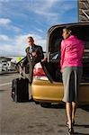 Businesswoman unloading her suitcase from a car trunk with a businessman standing in front of her