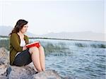 Frau sitzt an einem See in einem Journal schreiben