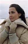 Young darkhaired Woman cuddling herself into the Collar of her winterly Jacket - Season - Gesture - Beach