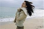 Young darkhaired Woman cuddling herself into the Collar of her winterly Jacket - Wind - Season - Gesture - Beach