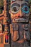 Nahaufnahme von einem Totem mit einer Frau, das Heraustreten aus einem Clan-Haus im Hintergrund von Totem Bight State Historical Park in Southeast Alaska