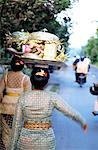 Woman ( women ) in embroidered dress with temple offerings walking down a road with motorcyclists in the distance . Ubud,Bali,Indonesia .