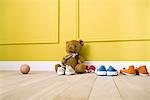 Teddy bear sitting on floor with several pairs of shoes, ball nearby