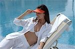 Woman dressed in white with an orange sun shade is sitting at the swimming pool