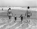 1970s FAMILY ON VACATION AT OCEAN BEACH HOLDING HANDS WALKING ON SAND IN SURF