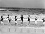 ANNÉES 1930 GROUPE 7 PERSONNES, MAIN DANS LA MAIN, À COURT DE SURF SUR LA PLAGE