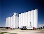 1970s WHITE GRAIN ELEVATORS TRUCKS PARKED IN FRONT OF SILOS AGAINST BLUE SKY