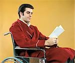 1970s MAN SITTING IN WHEELCHAIR WEARING RED BATHROBE READING GET WELL CARD
