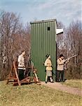 1960s TWO MEN ONE WOMAN WITH HUNTING RIFLES SHOTGUNS AMMO HUNTING GEAR STANDING BY TRAP ONE MAN AIMING GUN