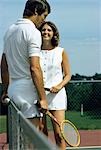 1970s SMILING COUPLE STANDING BY NET ON TENNIS COURT HOLDING RACKETS