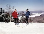 1960s COUPLE MAN WOMAN SKIERS TOP HILL WITH WINTER LAKE VISTA VIEW
