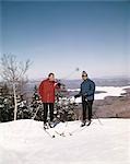 1960s COUPLE MAN WOMAN SKIERS TOP HILL WINTER LAKE VISTA VIEW