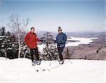 1960s COUPLE MAN WOMAN SKIERS STANDING TOP OF HILL WITH VISTA VIEW WINTER LAKE