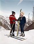 1960ER JAHRE LÄCHELND PAAR SPRECHEN AUF SKIPISTE TRAGEN SKI AUSRÜSTUNG