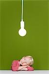 Little girl resting head on arms contemplating illuminated light bulb suspended overhead
