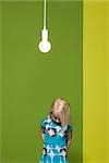 Little girl with hands behind back contemplatively looking down, illuminated light bulb suspended overhead