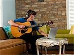 USA, Utah, Provo, junger Mann spielt Gitarre, mit Laptop im Wohnzimmer