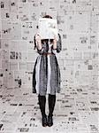 Young woman hiding face behind newspaper in newspapers covered room, studio shot