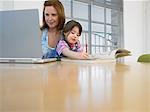 Girl (3-4) colouring in book beside mother using laptop, in home