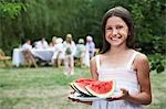 Girl (10-12) holding slices of watermelon in garden, family members in background
