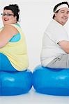 Overweight man and woman sitting back to back on exercise balls, portrait