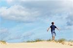 Young boy standing on sand dune in wind with arms outstretched, back view