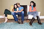 Third wheel sitting, arms crossed, on opposite end of sofa of hugging couple