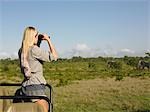 Young woman on safari, standing in jeep, looking through binoculars at elephants, back view
