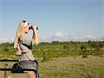 Young woman on safari, standing in jeep, looking through binoculars, side view