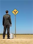 Businessman standing by briefcase, looking at road sign in desert, back view