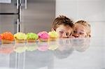 Young girl and boy peeking over counter at row of cupcakes, high section