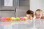 Young girl and boy looking over counter at row of cupcakes, in kitchen
