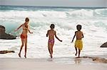 Three children (5-6, 7-9, 10-12) running on beach, back view