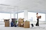 Office worker carrying carton near cartons and equipment on floor of empty office space