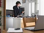 Homme debout dans la cuisine, la messagerie texte, ordinateur portable sur la table au premier plan